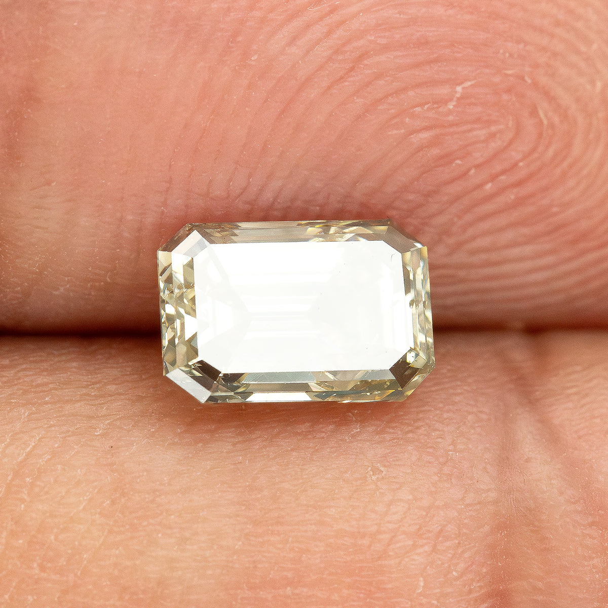 Emerald-cut diamond on fingertips at a highly reflective angle, with a broad white reflection across its face.