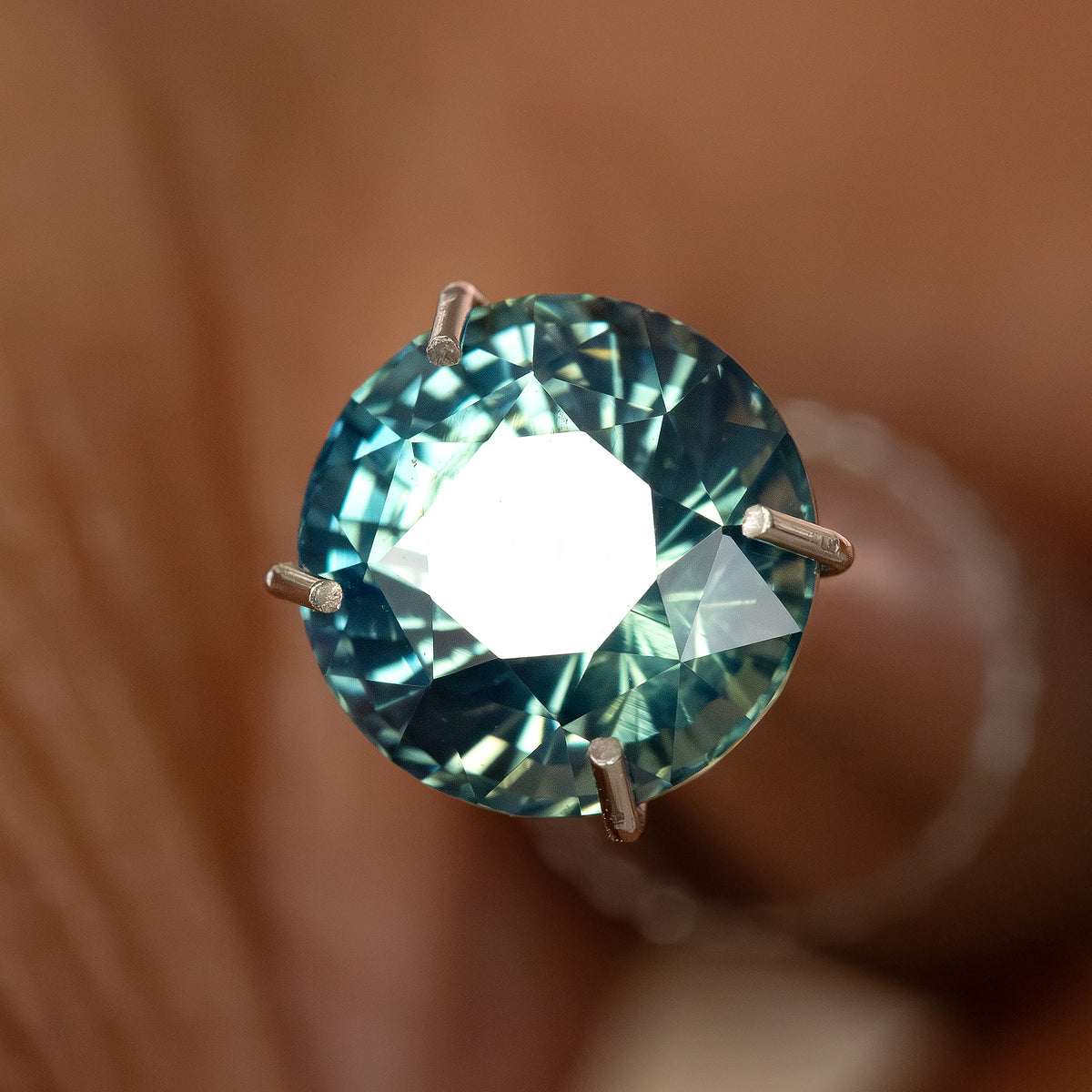 Round teal-blue and green sapphire in a temporary prong-style inspection holder, with a large white table reflection; holder not included.
