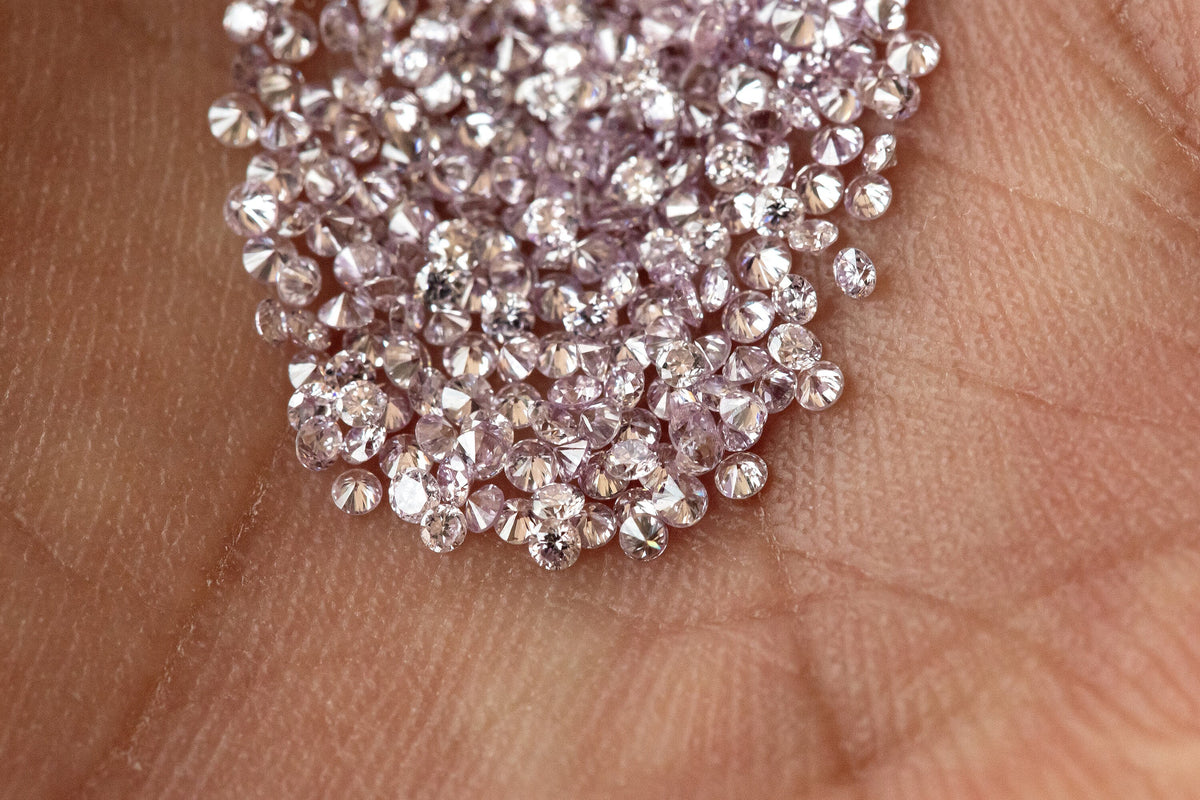 Representative purple-pink round diamond melee on an open palm for scale; one order is five stones.