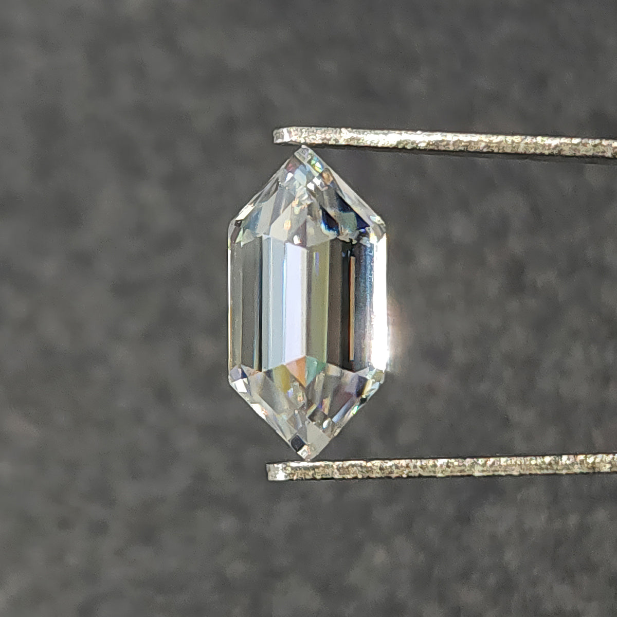Elongated hexagon step-cut colorless gemstone held vertically in tweezers against a dark grey background.