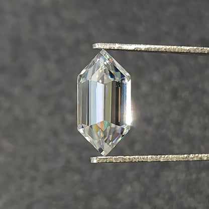 Elongated hexagon step-cut colorless gemstone held vertically in tweezers against a dark grey background.