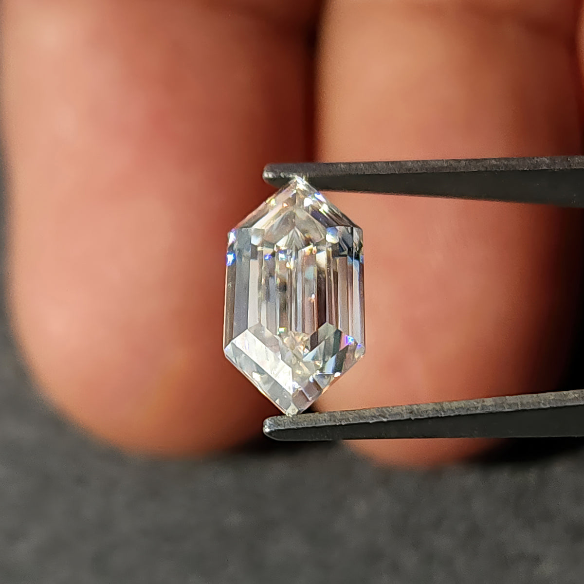 Colorless elongated hexagon step-cut gem held in tweezers with fingers blurred in the background.