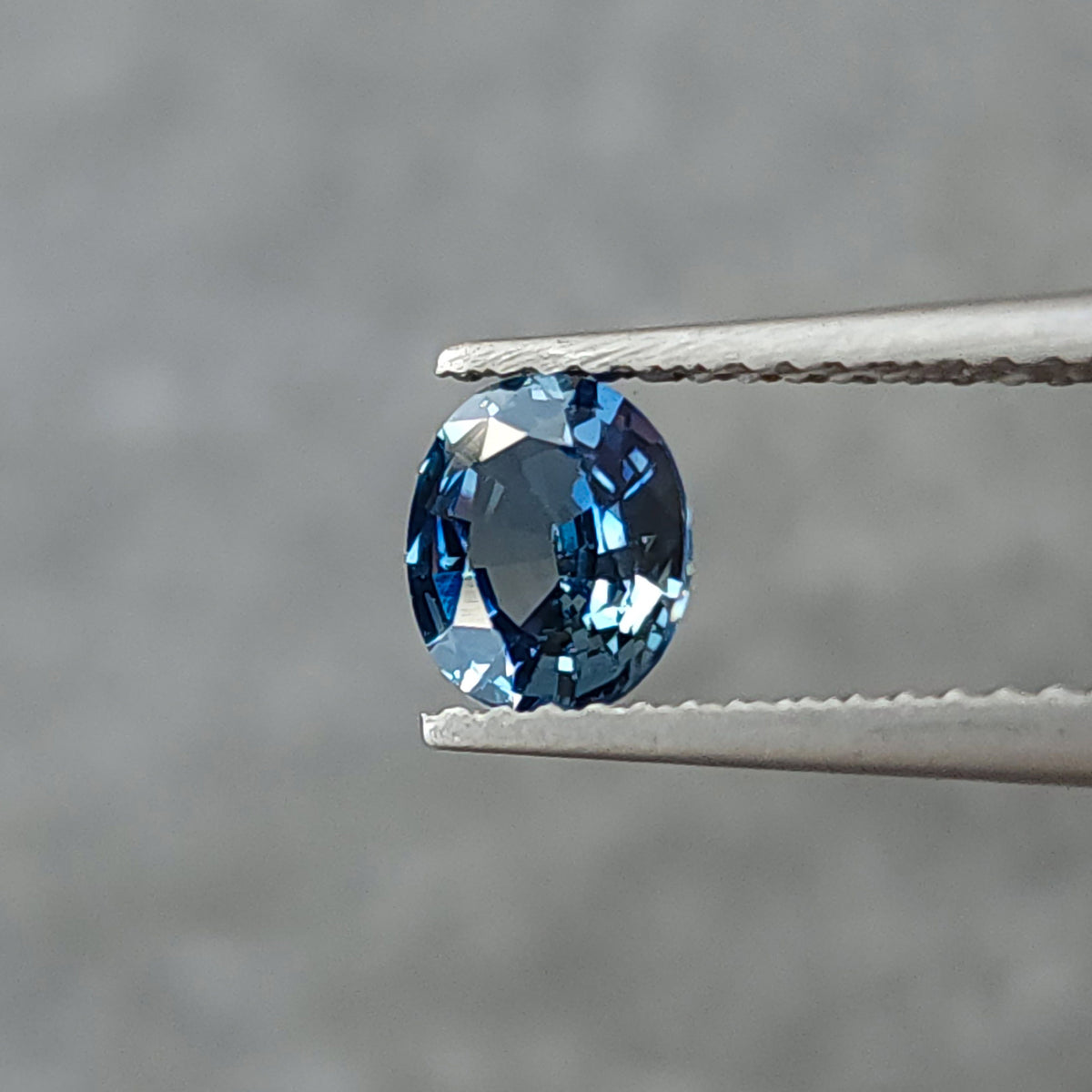 Blue-gray oval sapphire held upright face-up in tweezers, with blue rim reflections and a dark central area.