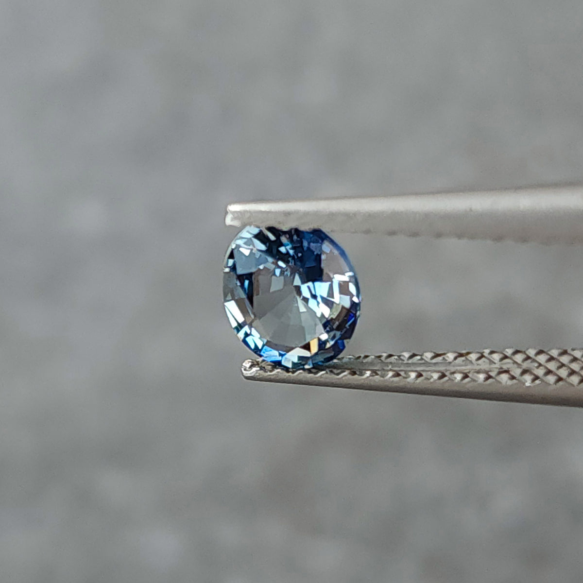 Blue-gray oval sapphire turned toward a shallow angle in tweezers, with gray and pale reflections.