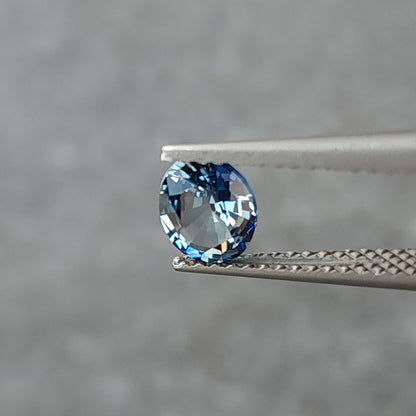 Blue-gray oval sapphire turned toward a shallow angle in tweezers, with gray and pale reflections.
