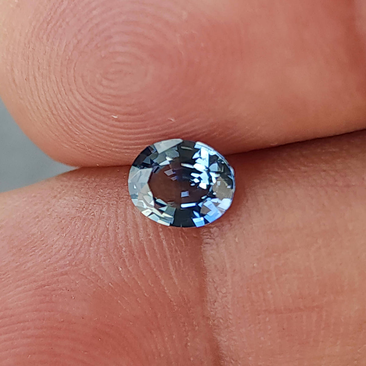 Oval blue-gray sapphire resting face-up between fingertips for scale, with its full outline visible.