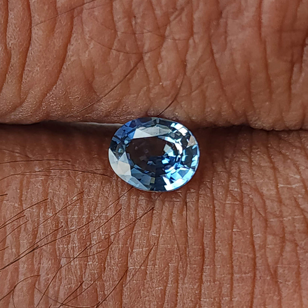 Blue-gray oval sapphire resting in a finger crease, viewed face-up against skin texture.