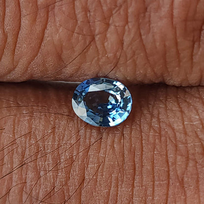 Blue-gray oval sapphire resting in a finger crease, viewed face-up against skin texture.