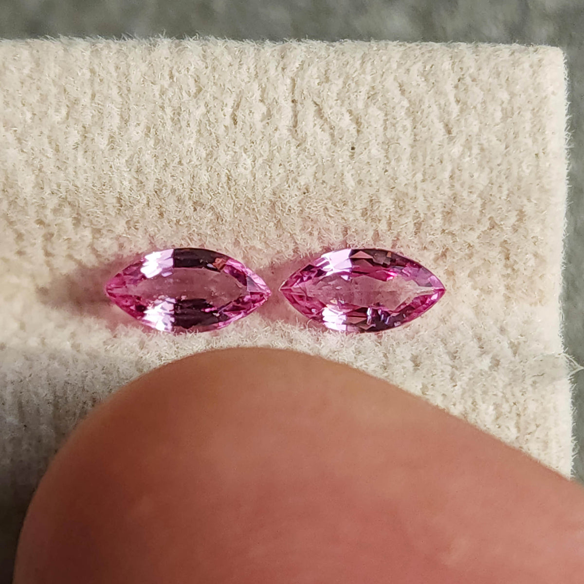 Two pink marquise sapphires on their cream pad beside a thumb for scale; they are not resting on the thumb.
