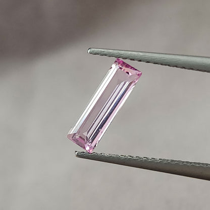 Pastel pink baguette sapphire held diagonally face-up in tweezers, showing its rectangular outline and step facets.
