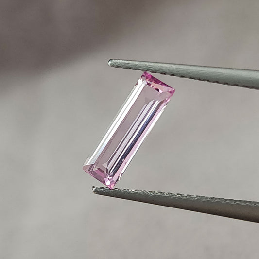 Pastel pink baguette sapphire held diagonally face-up in tweezers, showing its rectangular outline and step facets.