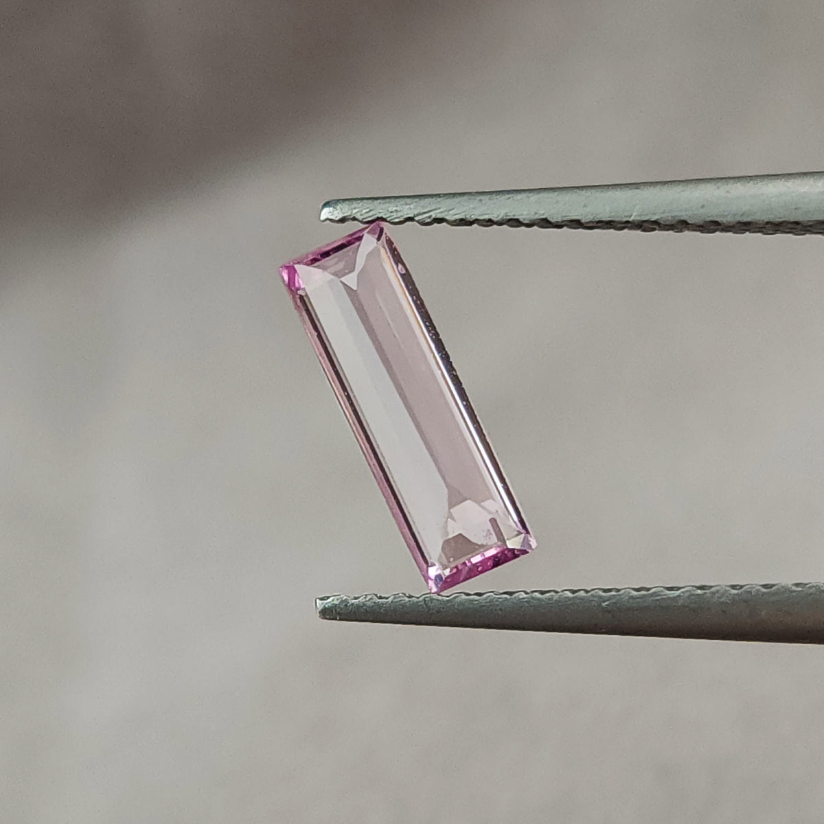 Pink baguette sapphire rotated toward an upright position in tweezers, with a broad pale central reflection.