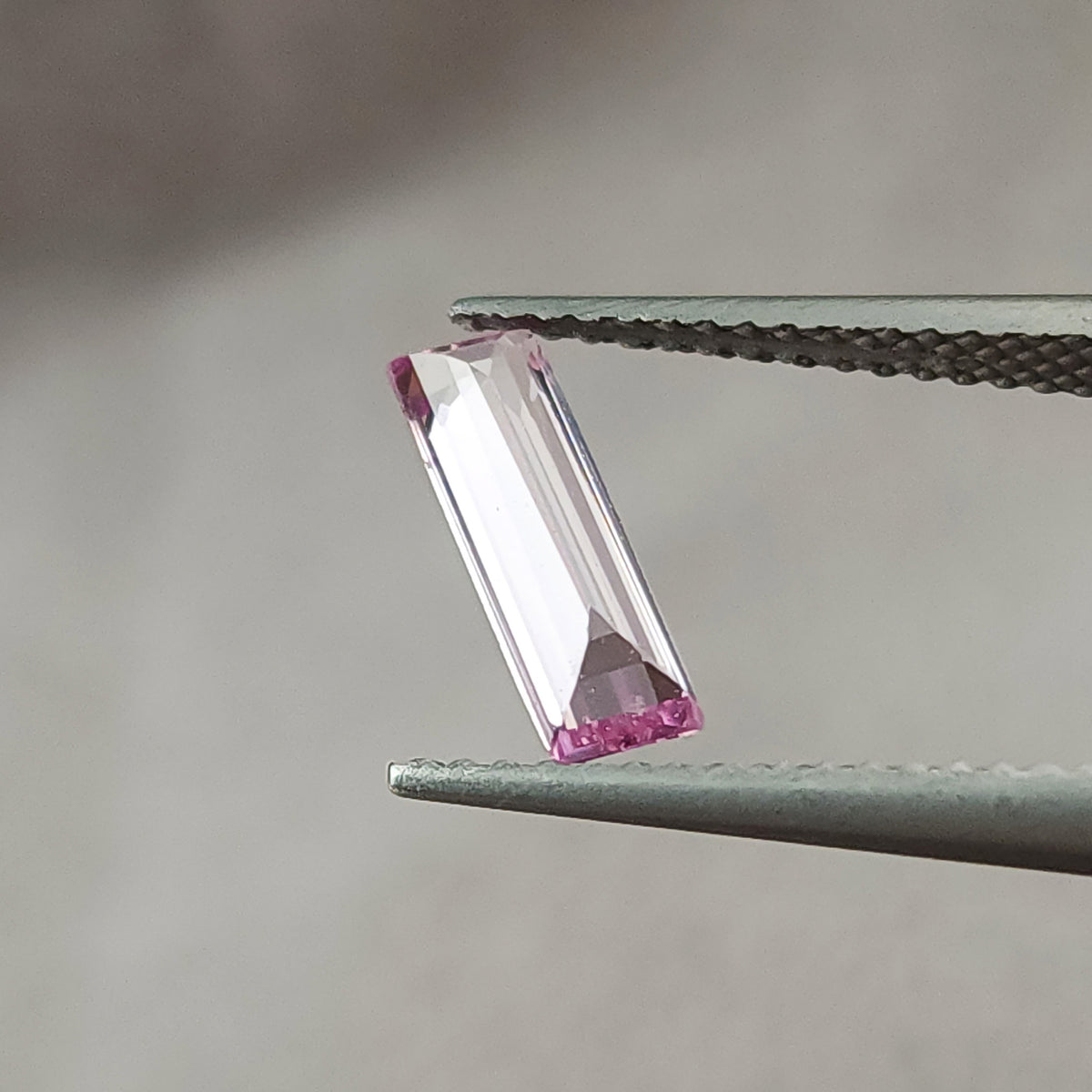 Tilted pink baguette sapphire in tweezers, with bright white reflections across the long facets.