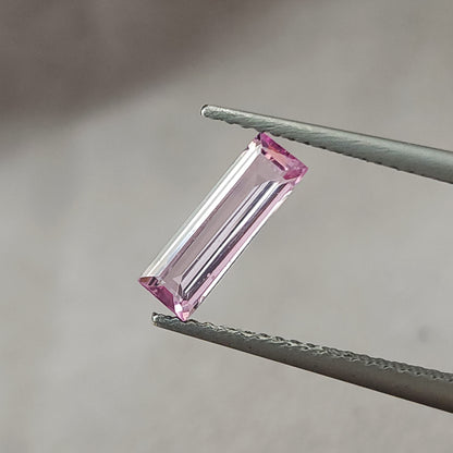 Alternate diagonal face-up view of the pink baguette sapphire between tweezers against a gray background.