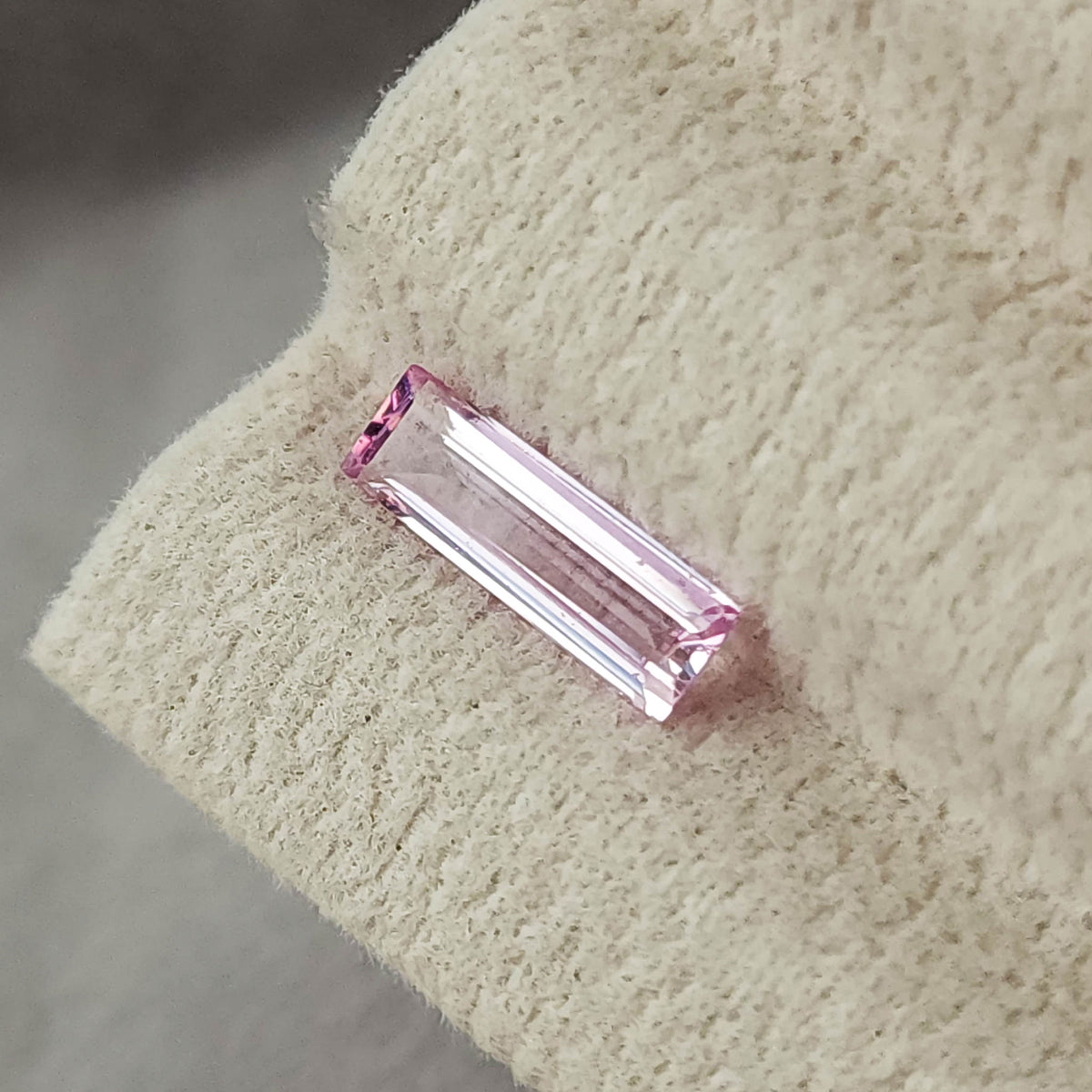 Pink baguette sapphire resting diagonally on a cream grooved display pad, viewed close-up.