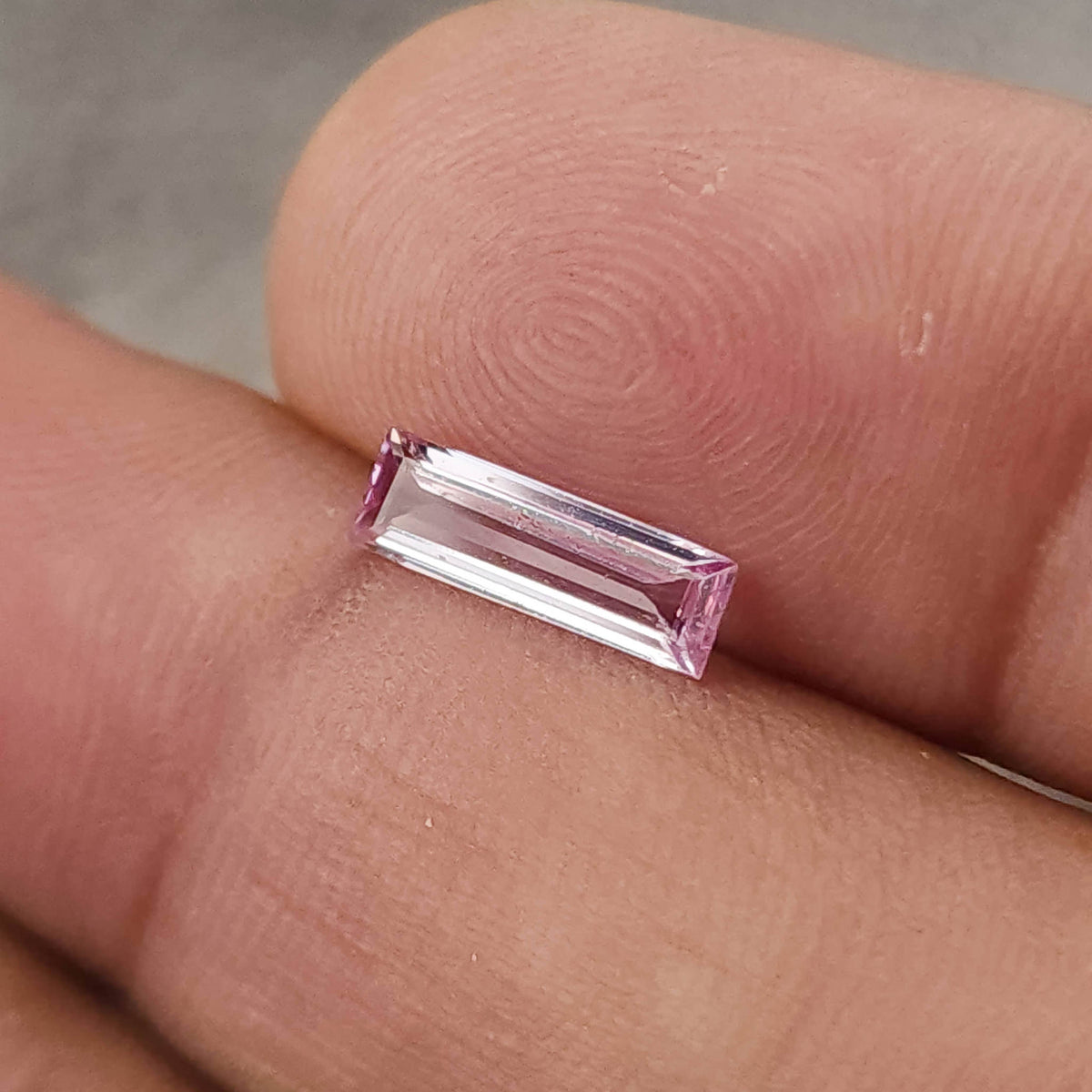 Pink baguette sapphire resting face-up between fingertips for scale, with skin ridges visible.