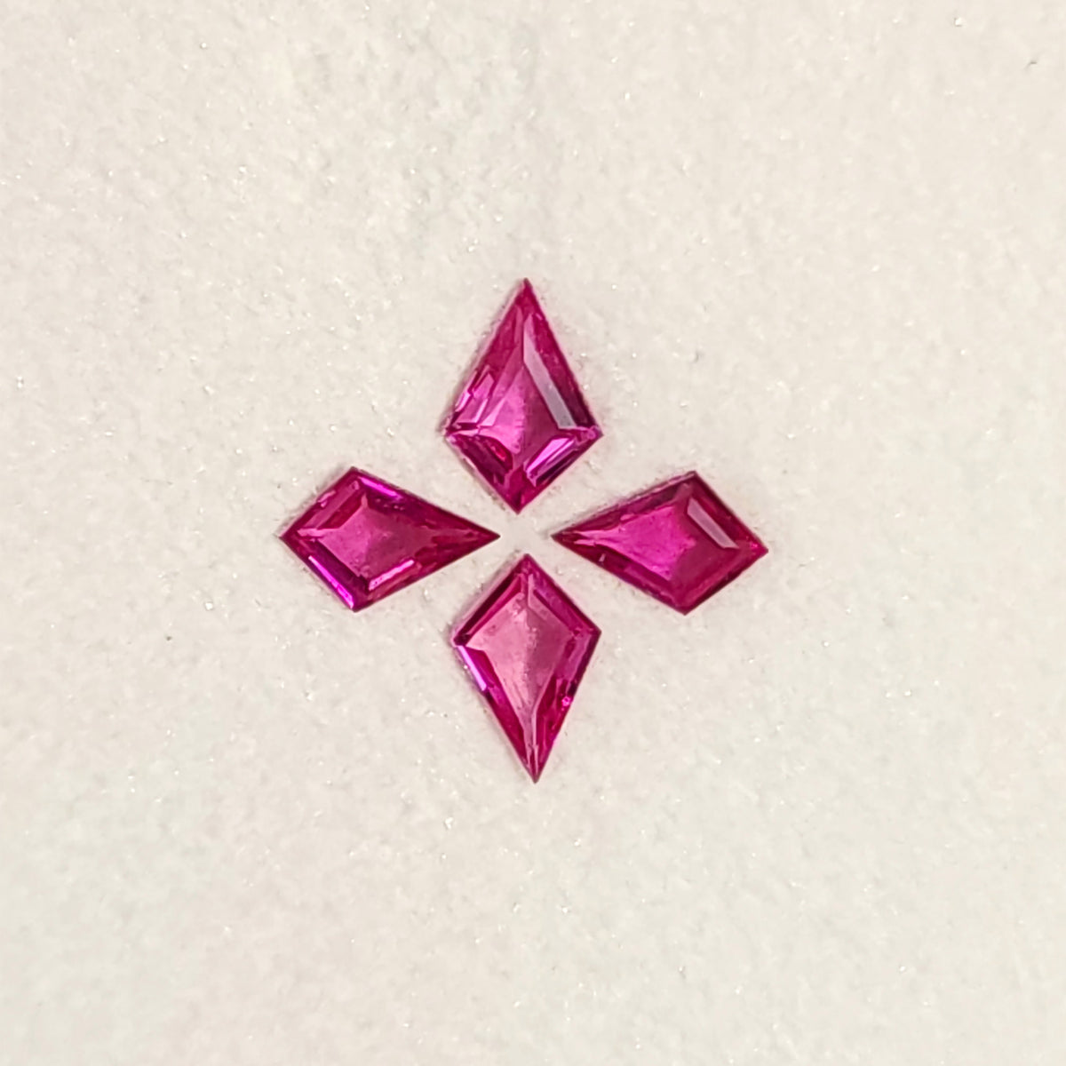 Close face-up view of all four fuchsia pink kite rubies, with their points facing outward on a white pad.