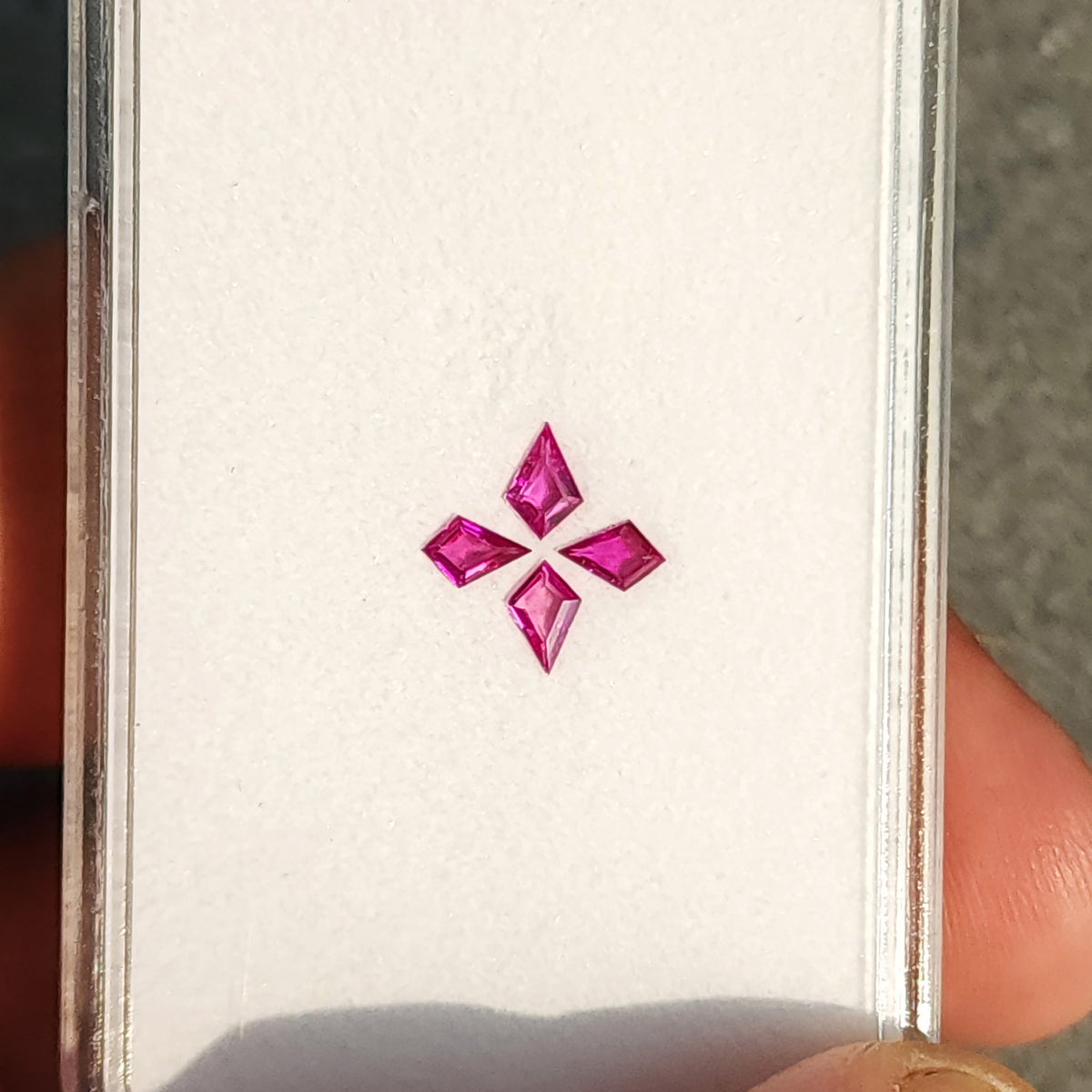 Wider upright tray view of the four pink kite rubies, with a fingertip visible at the lower right.