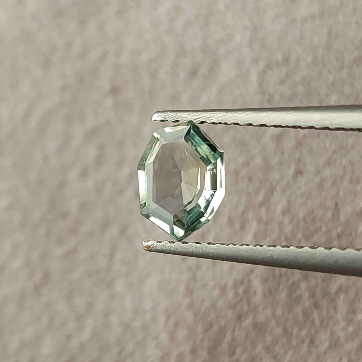 Mint green sapphire held upright in tweezers, viewed face-up against a textured pale surface.