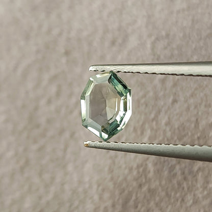 Mint green sapphire held upright in tweezers, viewed face-up against a textured pale surface.