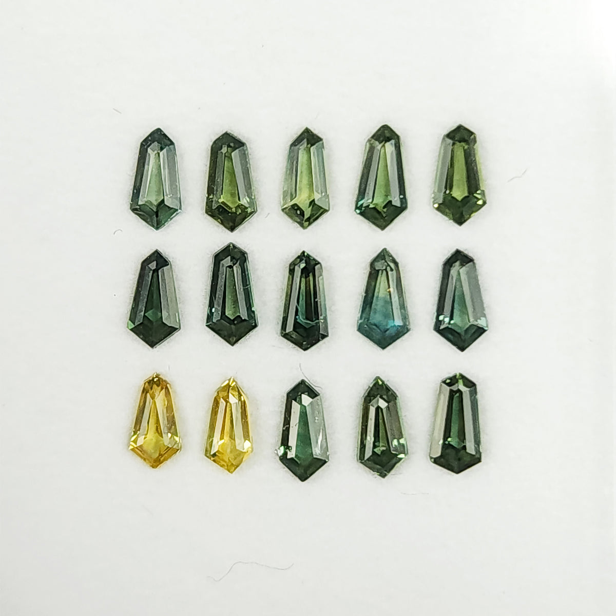 Fifteen green and yellow-green sapphires arranged face-up in three rows on a white tray.