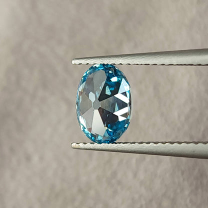 Blue oval diamond rotated in tweezers, showing its opposite broad reflective facet pattern.