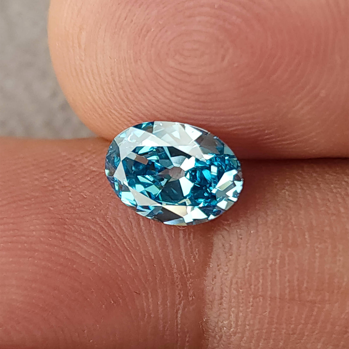 Blue oval diamond between fingertips for scale, with broad facets visible.