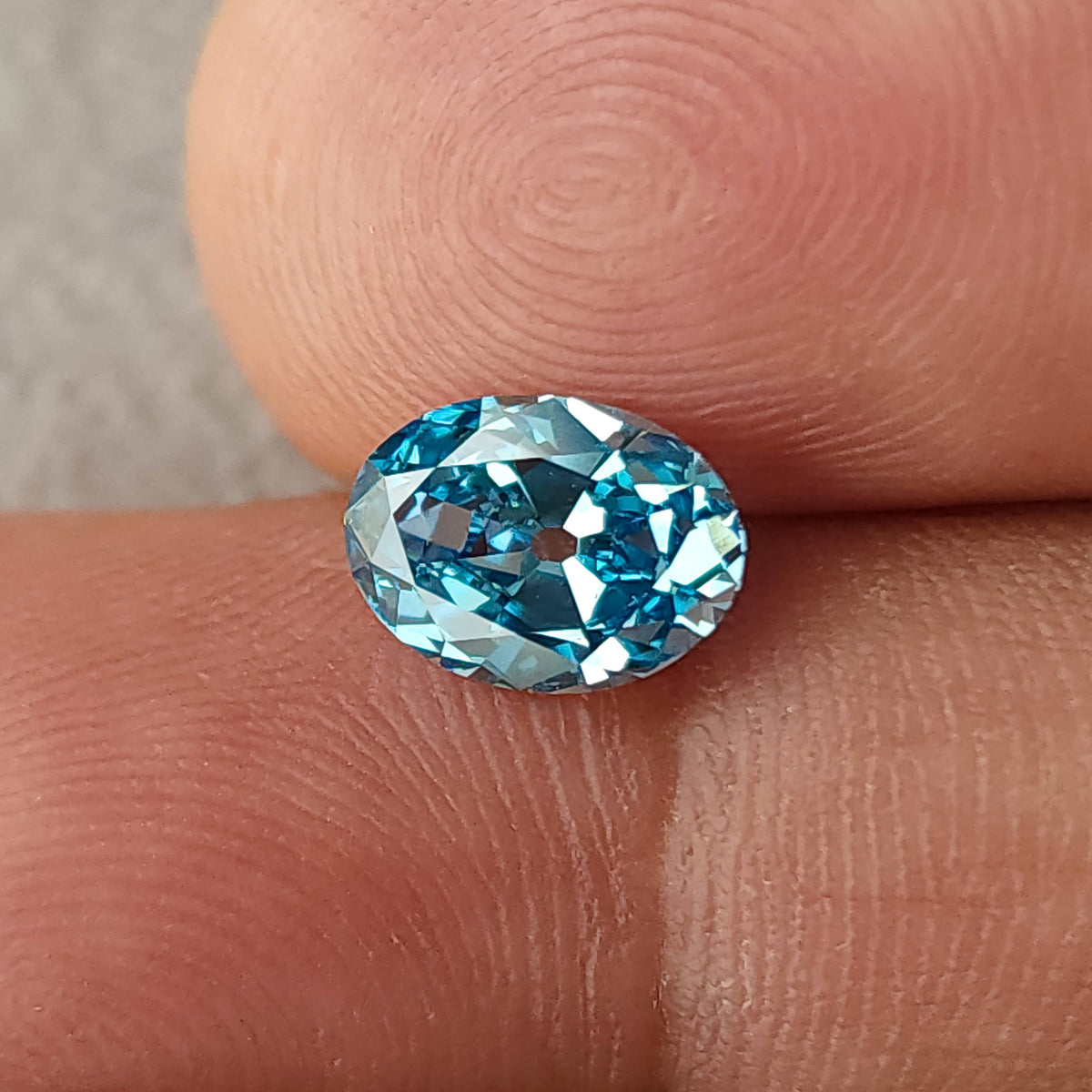Face-up blue oval diamond resting between two fingertips in a horizontal orientation.