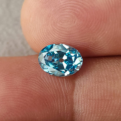 Face-up blue oval diamond resting between two fingertips in a horizontal orientation.