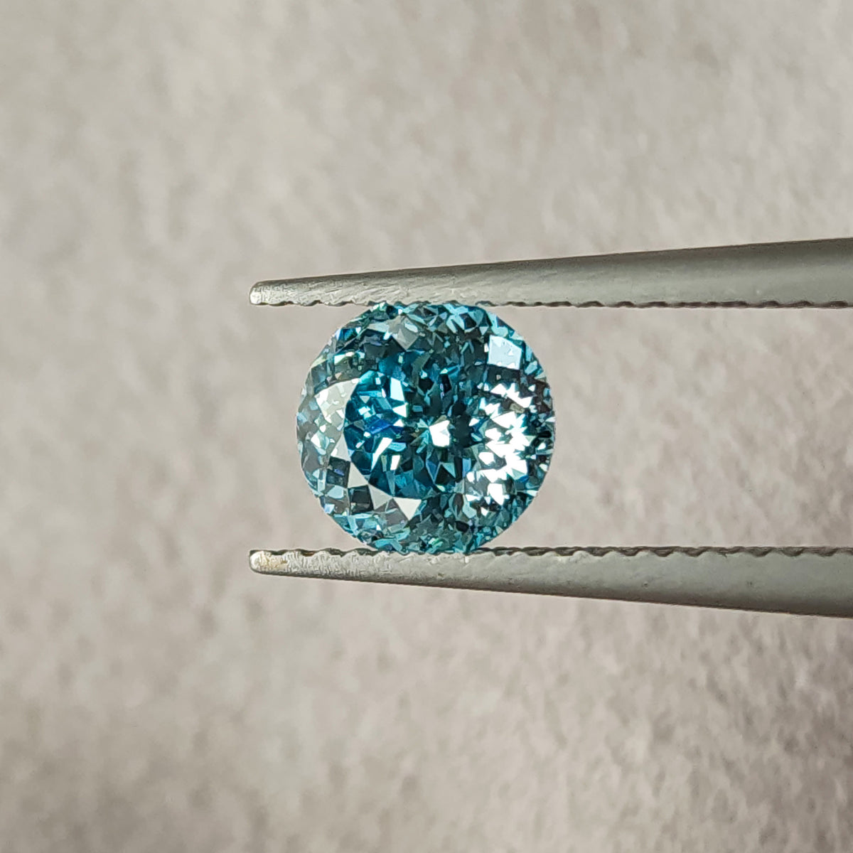 Face-up blue round lab-grown diamond held in metal tweezers over beige textured fabric, showing many small facets with blue and white reflections.