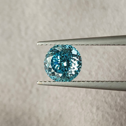 Face-up blue round lab-grown diamond held in metal tweezers over beige textured fabric, showing many small facets with blue and white reflections.
