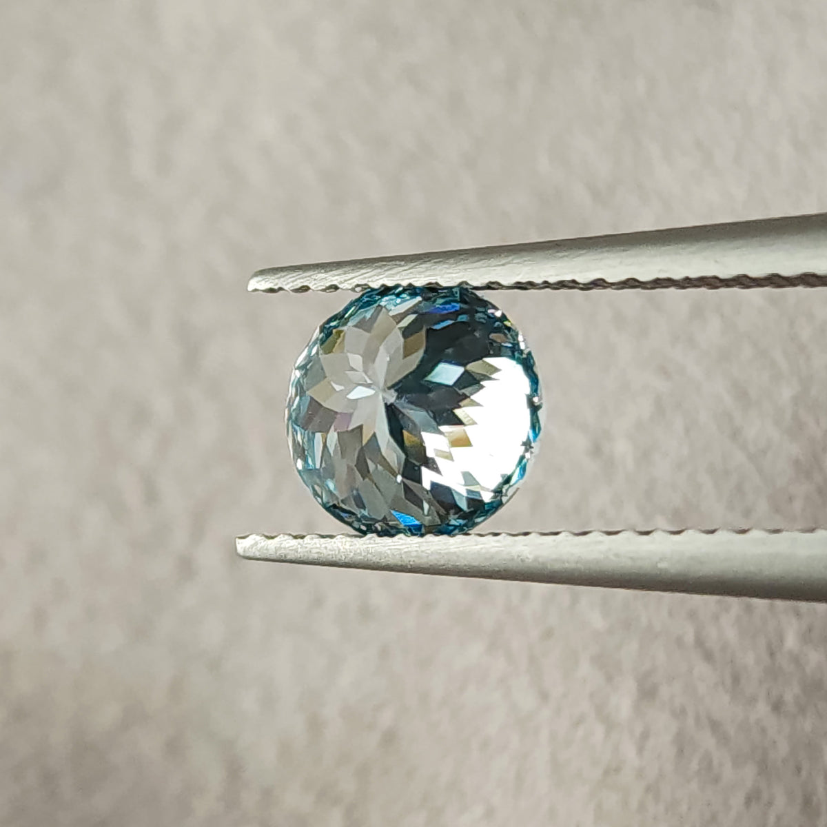 Reverse view of the blue round diamond held in tweezers, showing broad pavilion facets radiating from the center and pale reflections within a blue rim.