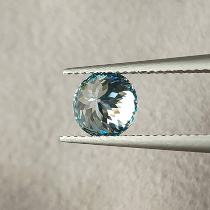 Reverse view of the blue round diamond held in tweezers, showing broad pavilion facets radiating from the center and pale reflections within a blue rim.