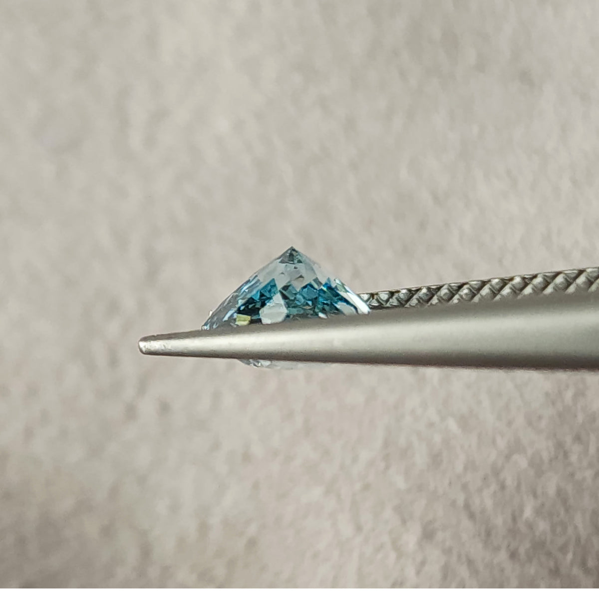 Side profile of the blue diamond held in metal tweezers, showing a pointed pavilion above the jaws; the front jaw partly covers the lower edge.