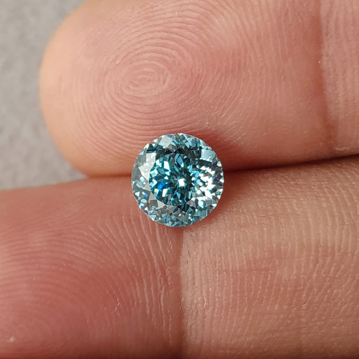 Face-up blue round lab-grown diamond resting on fingertips; fingerprint ridges provide scale beside the blue and white facet reflections.