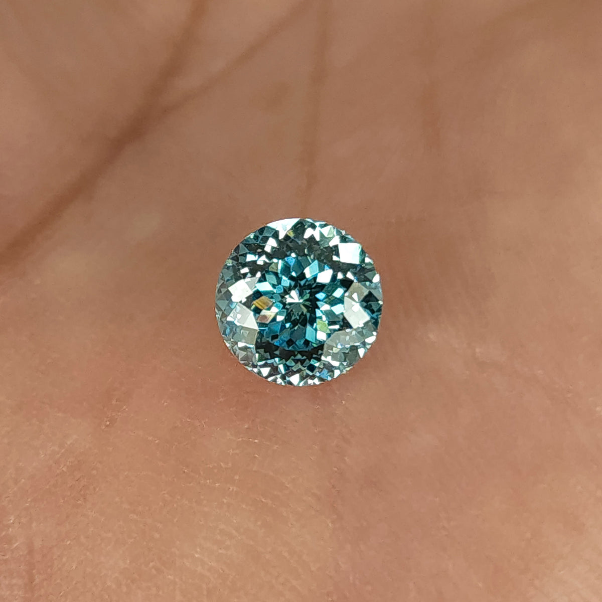 Blue round diamond resting face-up on skin, with a star-like center, numerous small facets and bright white reflections around the edge.