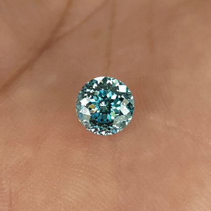 Blue round diamond resting face-up on skin, with a star-like center, numerous small facets and bright white reflections around the edge.
