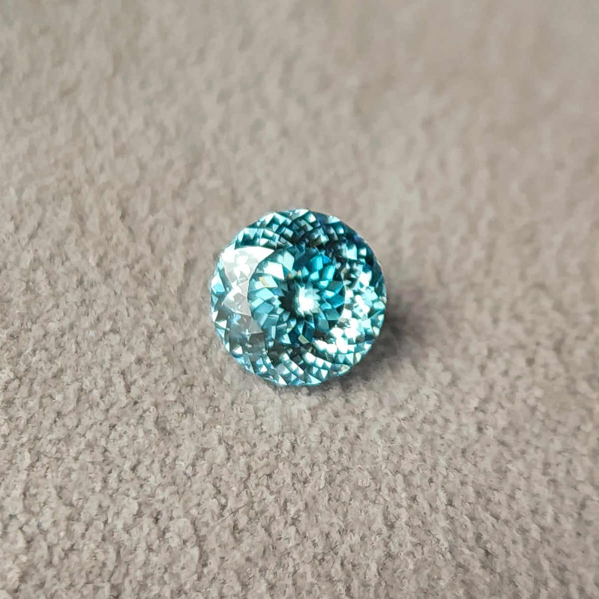 Blue round diamond on gray textured fabric, viewed almost face-up with concentric rows of facets and bright white reflections against the blue center.