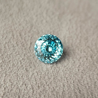 Blue round diamond on gray textured fabric, viewed almost face-up with concentric rows of facets and bright white reflections against the blue center.
