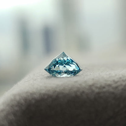 Side view of the blue round diamond on gray textured fabric, with its pointed pavilion facing upward and rows of blue and pale facets visible.