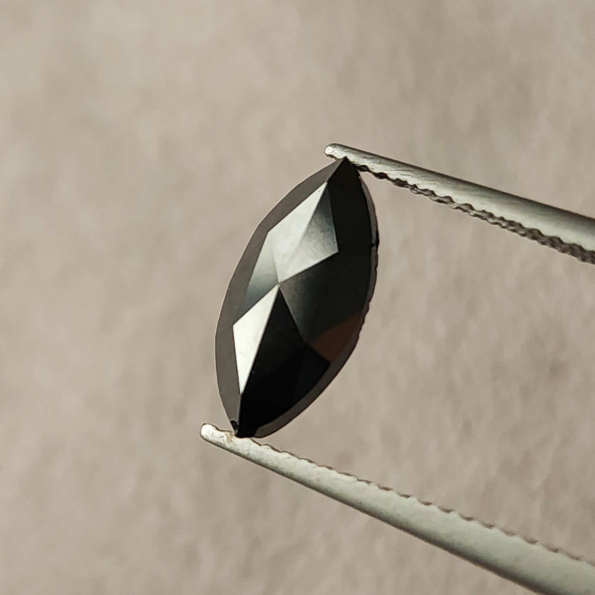 Black marquise rose-cut diamond held face-up in tweezers, with contrasting black and pale triangular facet reflections.