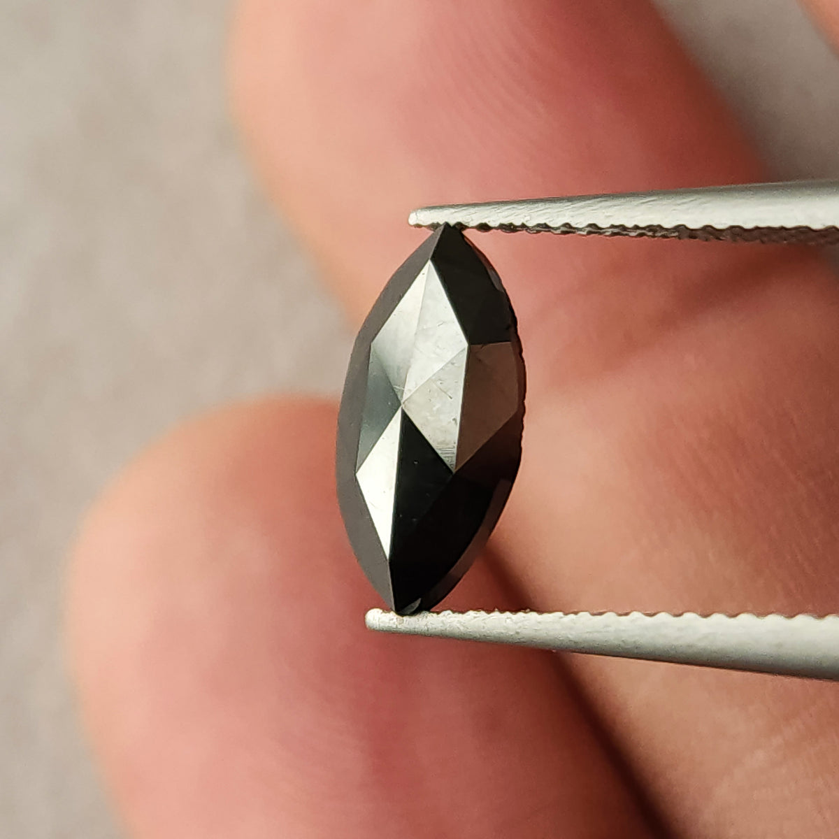 Black marquise diamond held upright in tweezers near fingers, showing its pointed outline and scale.