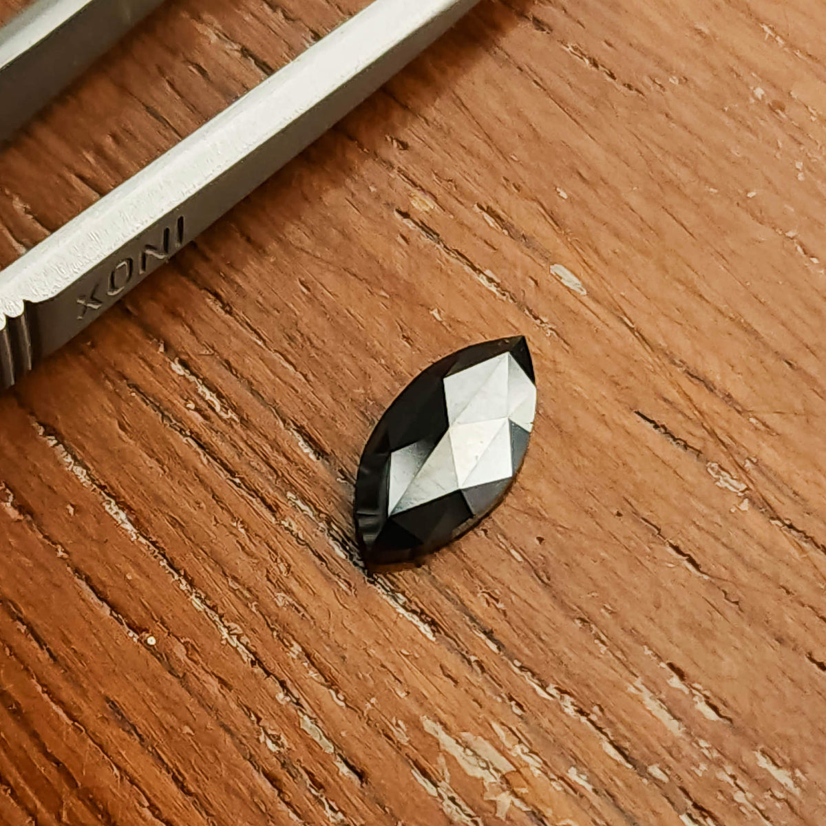 Black marquise diamond lying face-up on a wooden surface beside tweezers.