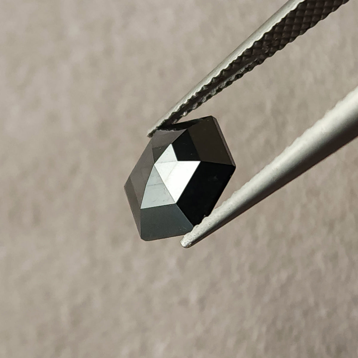 Tilted side view of the natural black hexagon diamond in tweezers, showing its angular outline and raised crown.