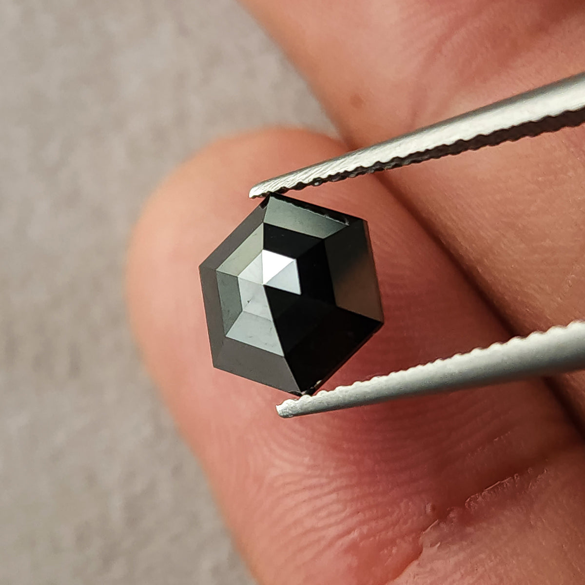 Face-up view of the natural black hexagon diamond held in tweezers beside a fingertip for scale.