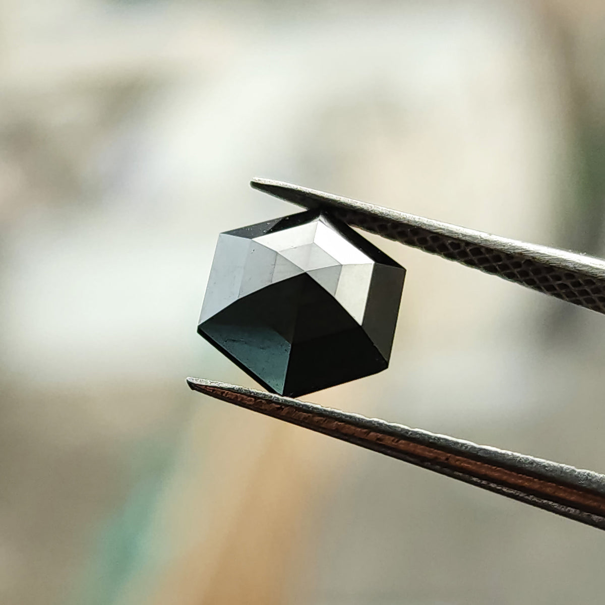 Angled face-up view of the rose-cut black hexagon diamond in tweezers, with broad silver reflections across the crown facets.