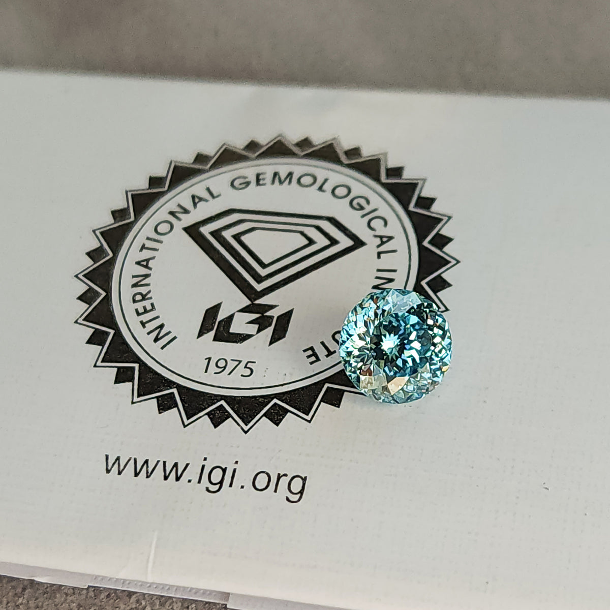 Blue round diamond placed on the printed IGI logo of a laboratory report, with the circular logo and website visible beside the stone.