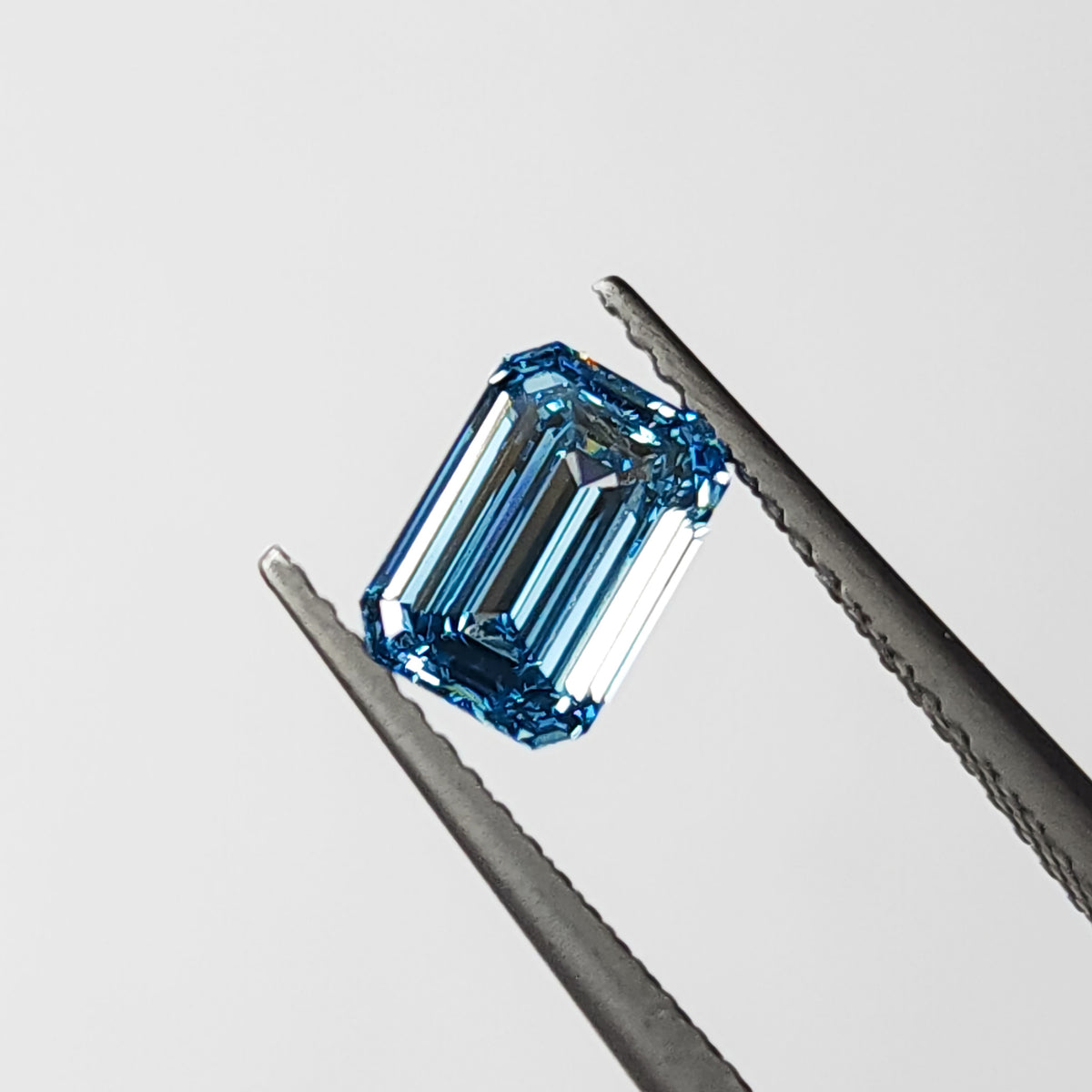 Blue lab-grown emerald-cut diamond held face-up in tweezers, showing its rectangular table, clipped corners and step facets.
