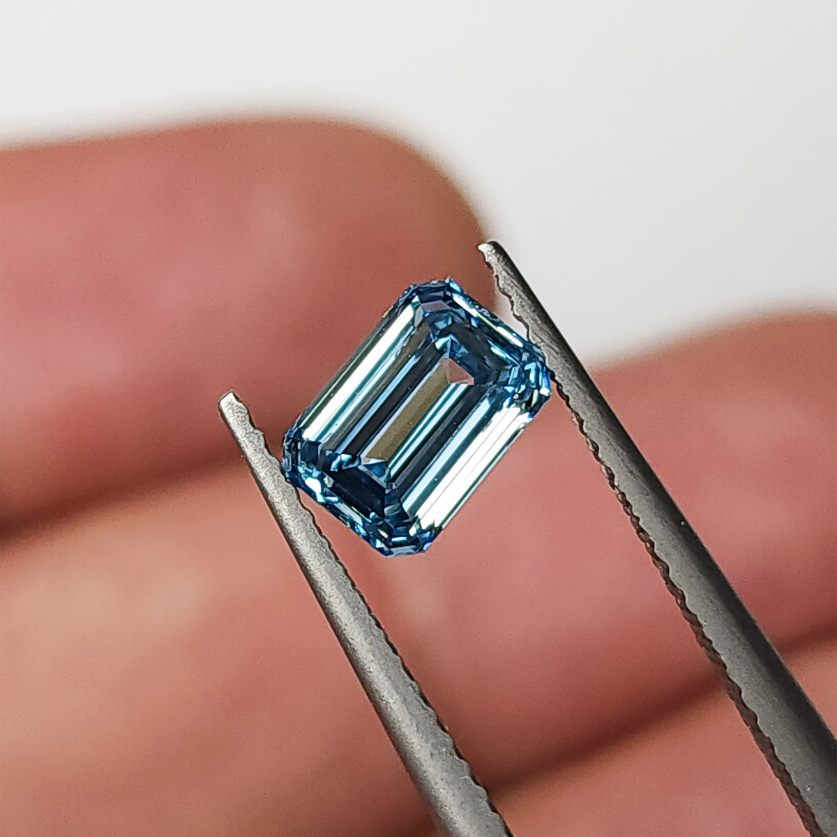 Blue lab-grown diamond held face-up in tweezers against a finger background for scale context.