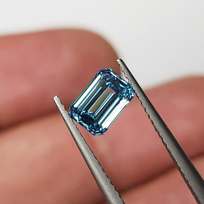 Blue lab-grown diamond held face-up in tweezers against a finger background for scale context.